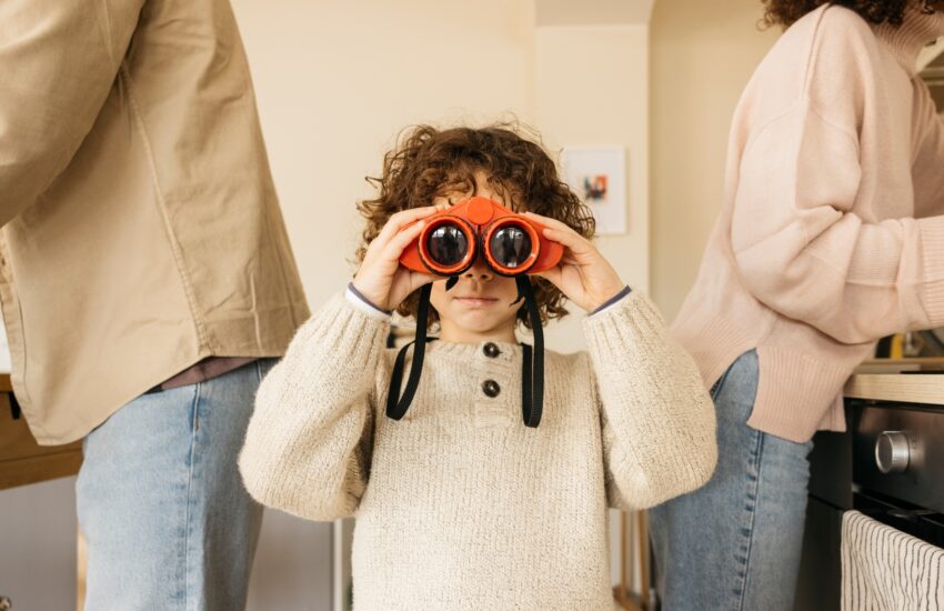 Child playing in a kitchen with toy binoculars amidst his parents.