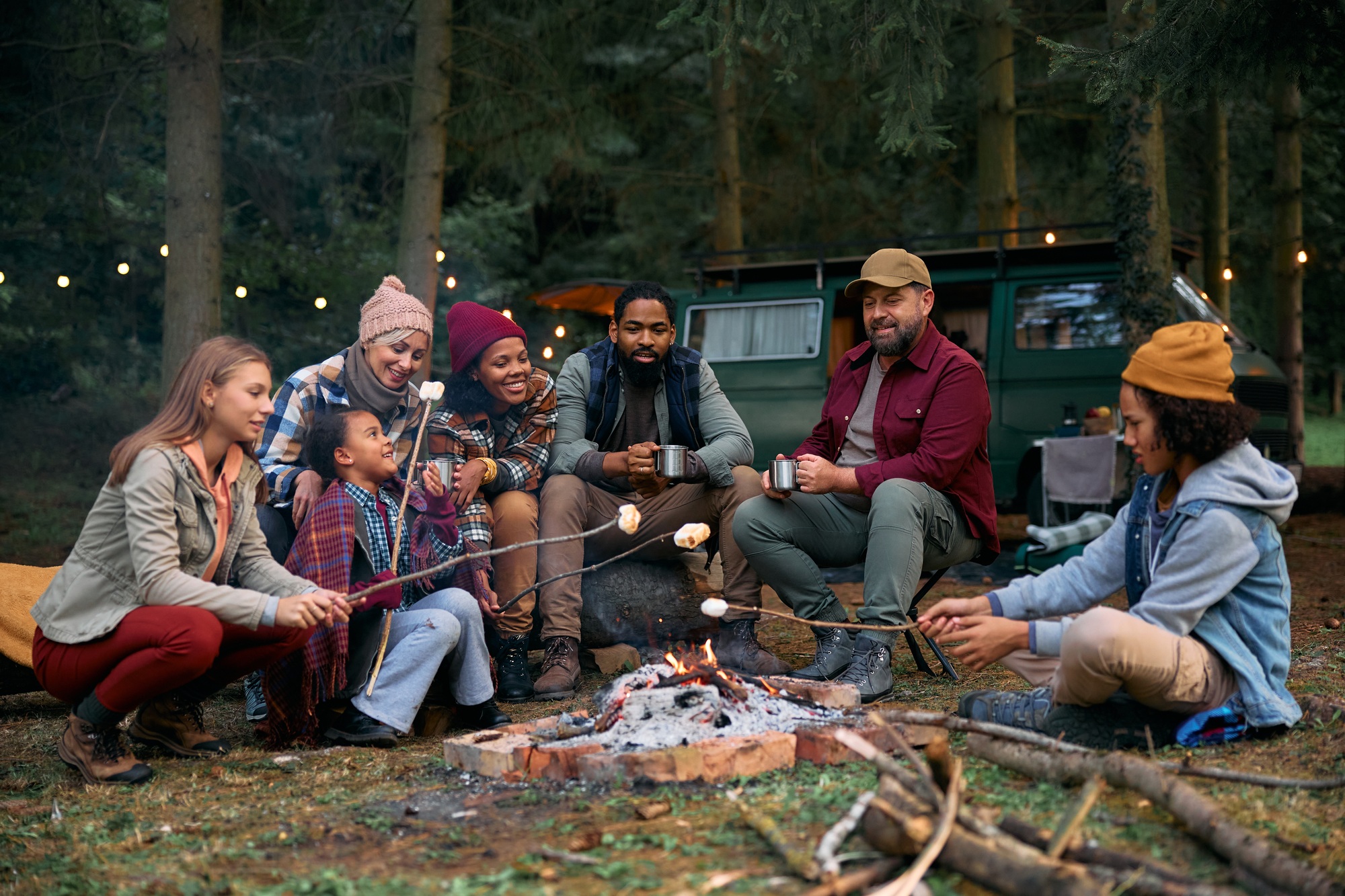 Multiracial families roasting marshmallows while gathering around campfire in the woods.